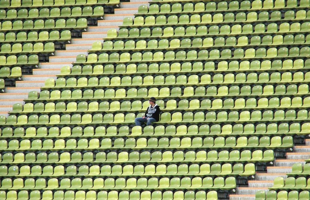 Stadiona tribīnes ar vairākiem simtiem krēslu zaļā krāsā. Vienā no krēsliem sēž vīrietis. 