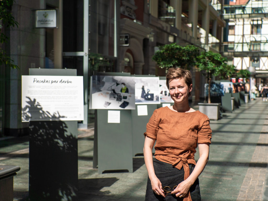 Māksliniece Zane Veldre stāv "Galerijas Centrs" galvenajā ielā pie pasakām par darbu veltītās izstādes galvenā stenda, kurā aprakstīta izstādes ideja. Fonā viņai ir redzami vairāki citi stendi, uz kuriem izvietotas pasakas un to ilustrācijas.