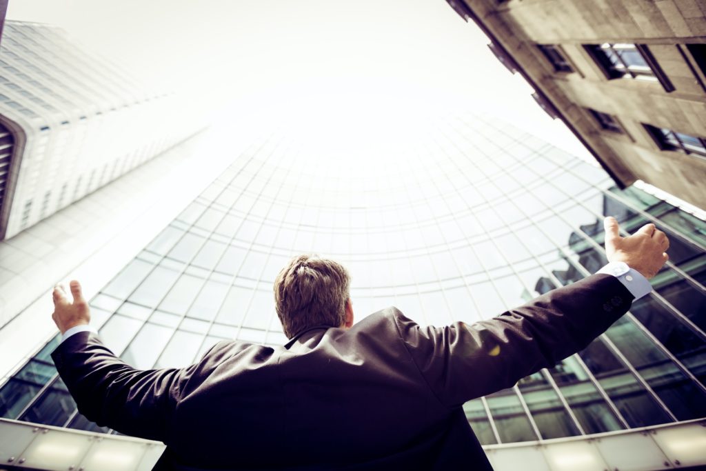 Vīrietis ar izplestām rokām stāv lielas, stilklotas biroja ēkas pakājē un skatās uz augšu. Blakus redzamas ir citas ēkas.