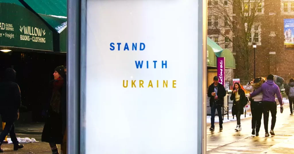 Vides reklāmas stends Toronto pilsētā, ASV, kurā uz baltas lapas Ukrainas karoga krāsās (dzeltenā un zilā) ir pausts atbalsts Ukrainai tās karā ar Krieviju: "Stand with Ukraine". Šī stenda fonā pa ielu mierīgi pastaigājas cilvēki.