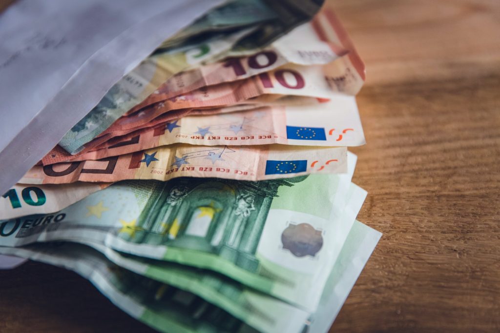 Uz galda stāv atvērta aploksne, no kuras daļēji izslīdējušas vairākas 5,10 un 50 eiro banknotes.