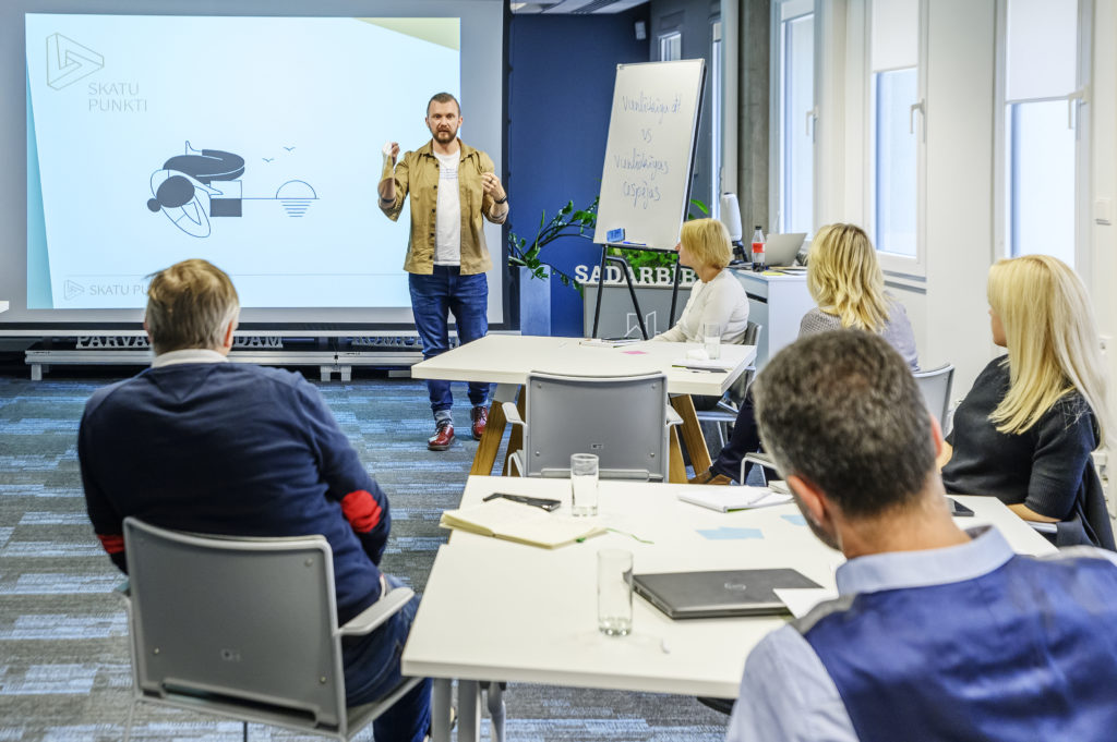 Foto mirklis no mācību programmas "Skatu Punkti" grupas, kas pulcēja dažādu organizāciju augstākā līmeņa vadītājus. Fonā ir šīs mācību programmas pasniedzējs Kaspars Zālītis, kurš uzrunā dalībniekus. Viņš stāv pie tāfeles, uz kuras rakstīts teksts "vienlīdzība vs. vienlīdzīgas iespējas".