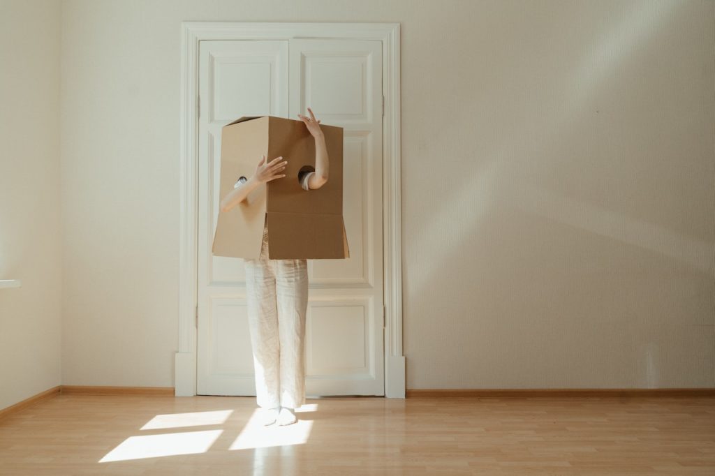 Tukšā telpā uz baltu durvju fona stāv cilvēks baltās biksēs. Viņa ķermeņa augšdaļu sedz kartona kaste, kurai izgriezti divi caurumi rokām.